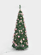 A decorated artificial Christmas tree with green branches, pink ornaments, and light pink ribbon bows, shown against a plain light gray studio background.