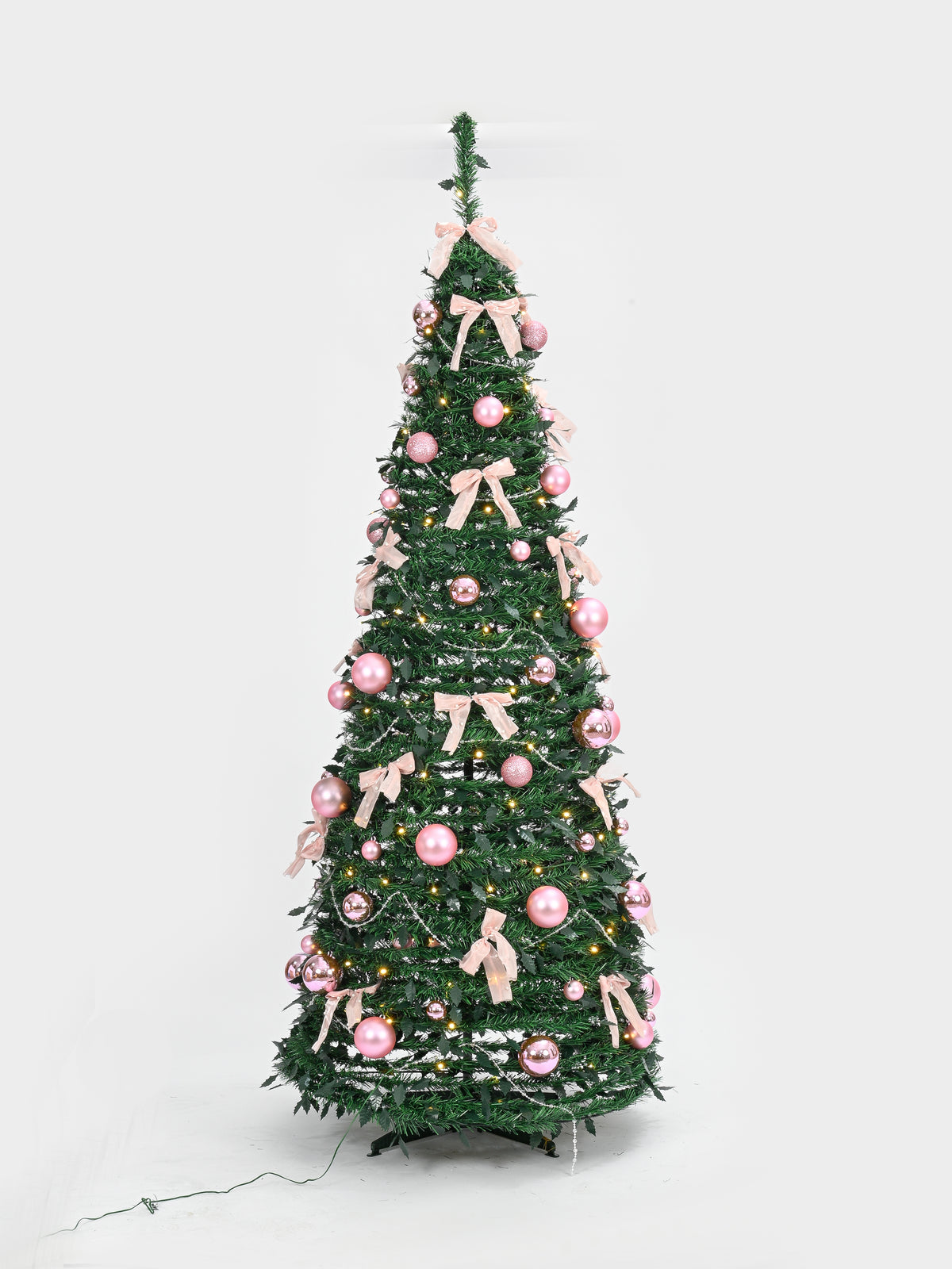 A decorated artificial Christmas tree with green branches, pink ornaments, and light pink ribbon bows, shown against a plain light gray studio background.